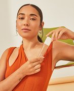 Photo of person in an orange shirt with their dark hair pulled into a slicked back ponytail. They are leaning against a green chair and holding the Lancôme Teint Idole Ultra Wear Care and Glow Foundation.