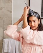 Person with long dark hair in a pale pink robe coloring their roots at home. 