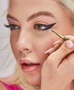 Person with light blonde hair and long nails using an eyeliner brush to apply a black winged eyeliner look