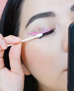 Picture of a person wearing colorful eyeliner and holding a cotton swab to their eyelashes.