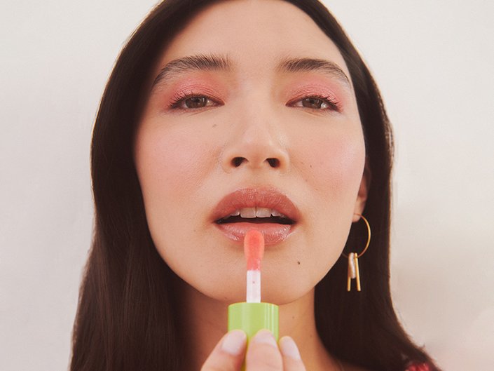 A model with dark hair and pastel pink eyeshadow applying a pale pink shade of the NYX Professional Makeup Fat Oil Lip Drip to their lips.  